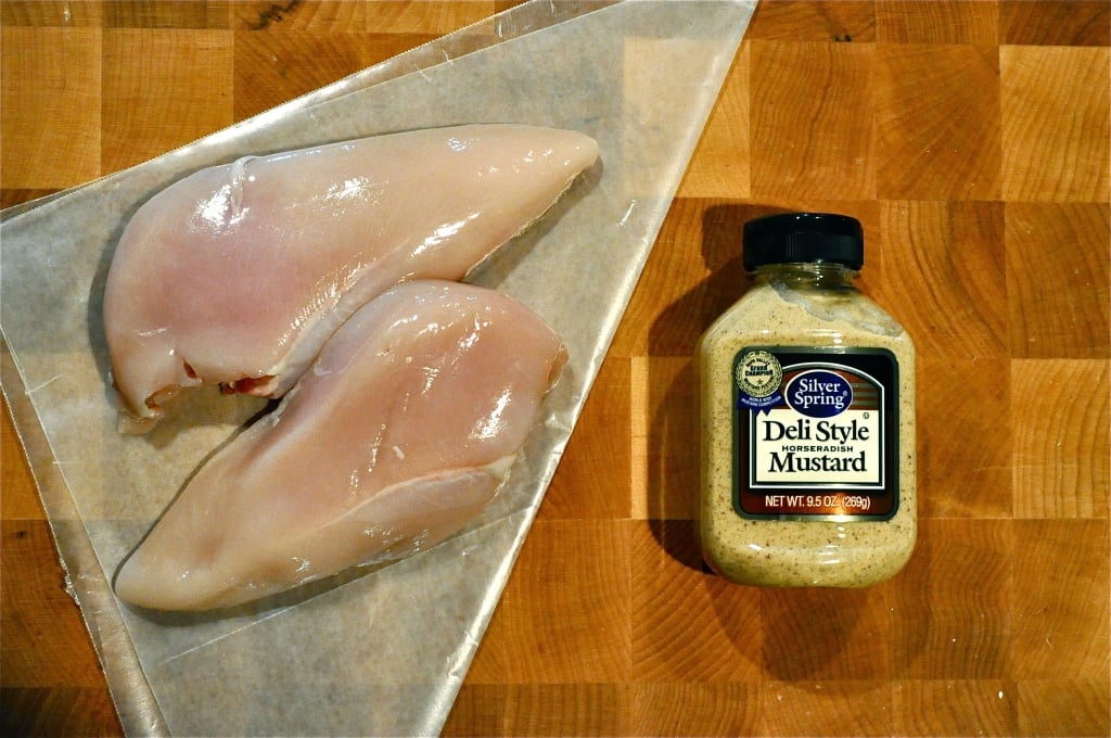 overhead view of two raw chicken breasts on top of wax paper on a wooden cutting board with deli style mustard next to it