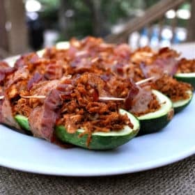 bacon wrapped stuffed zucchinis on a white plate on top of a burlap covered table