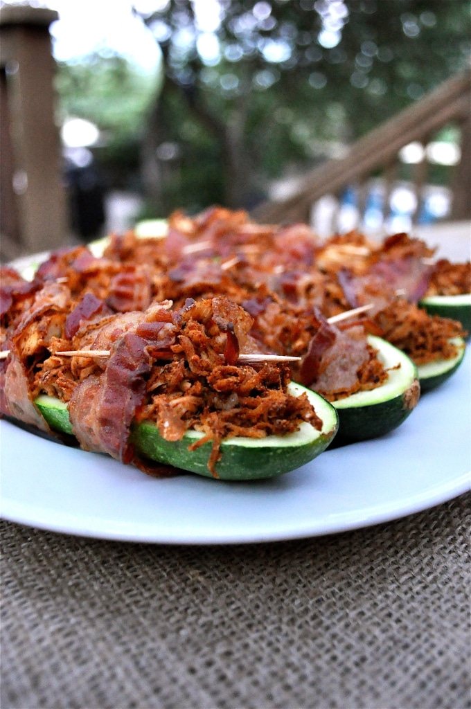 bacon wrapped stuffed zucchinis on a white plate on top of a burlap covered table