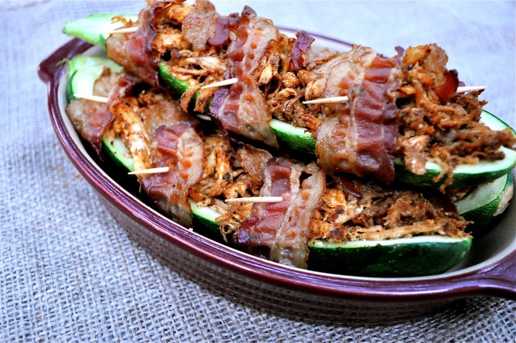 bacon wrapped stuffed zucchinis in a red casserole dish on top of a burlap covered table