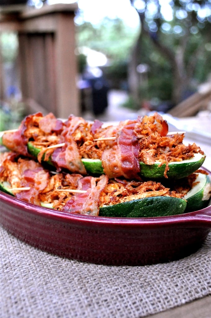 bacon wrapped stuffed zucchinis in a red casserole dish on top of a burlap covered table