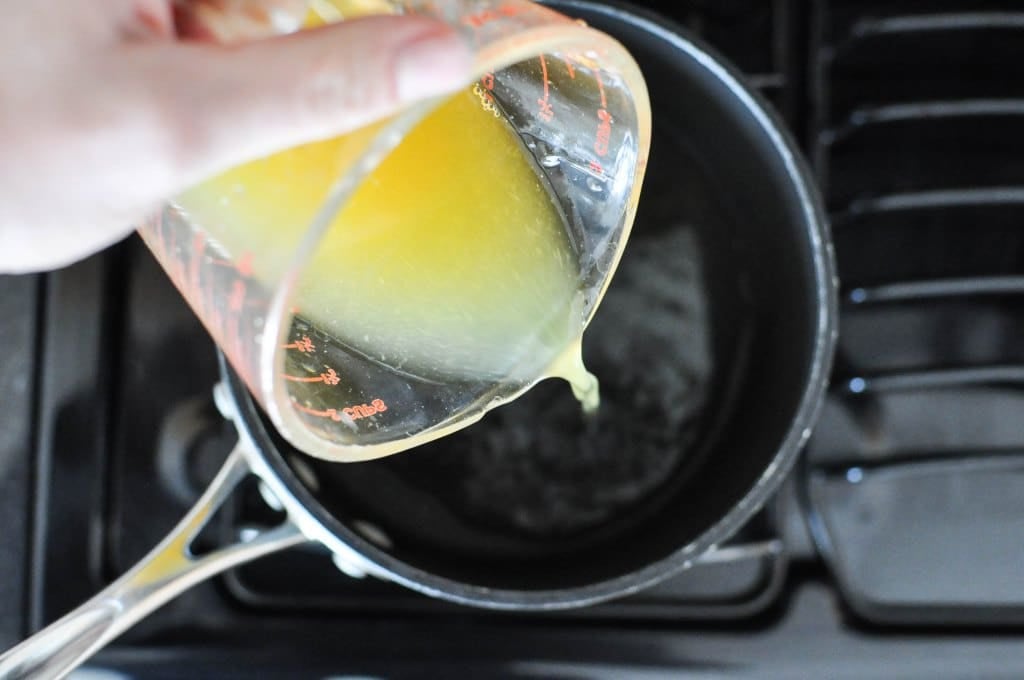 glass measuring cup of orange juice being poured into a black saucepan on a gas stovetop