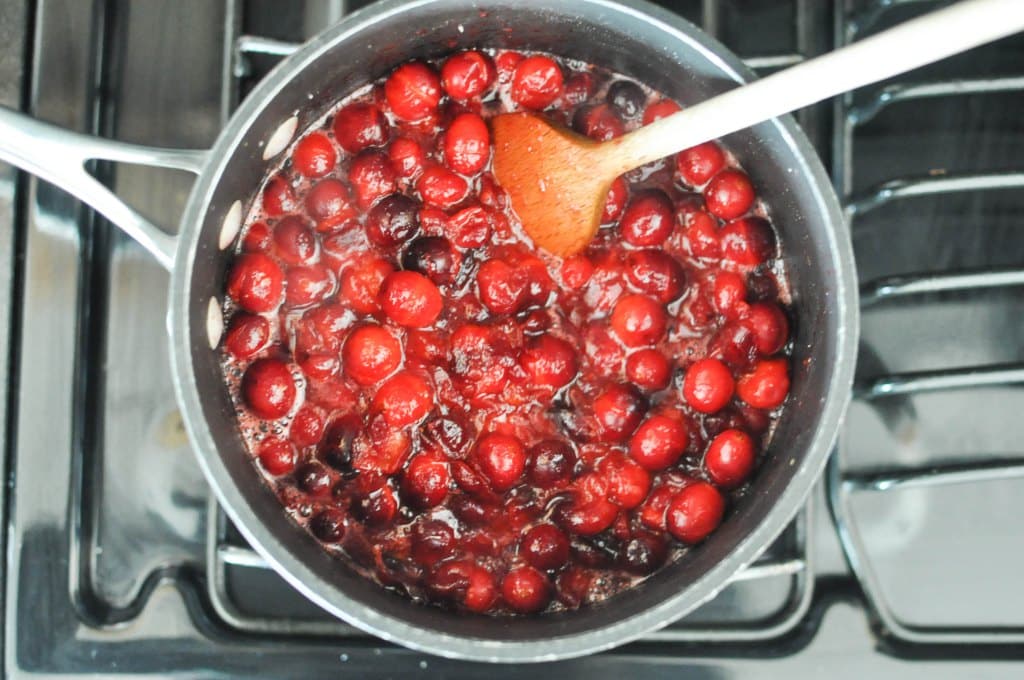bubbling cranberry sauce being stirred by a wooden spoon in a grey saucepan on a stovetop