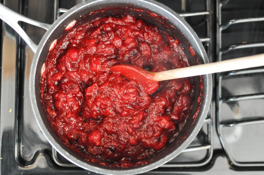 cranberry sauce in a grey saucepan on a stovetop being stirred by a wooden spoon