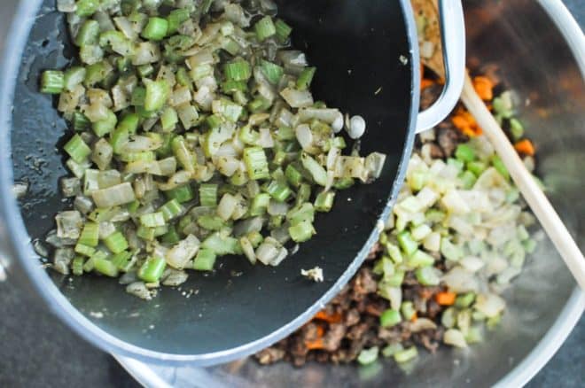 cooked onion and celery being added to a stainless steel bowl filled with browned sausage and sweet potatoes