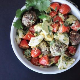 overhead view of a white bowl filled with chopped hard boiled eggs, cherry tomatoes, avocados, cilantro and sausage meatballs on a dark gray surface