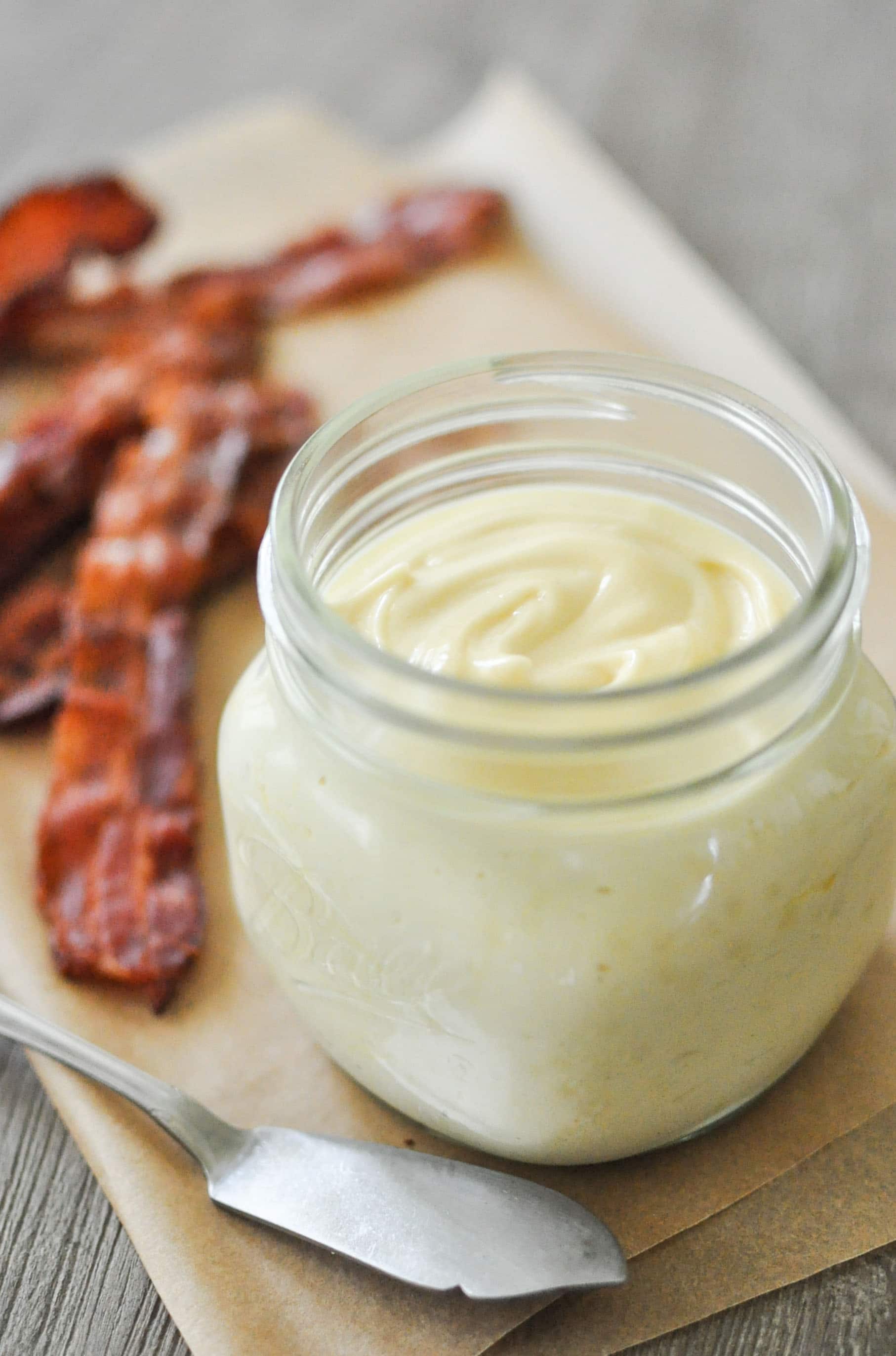a glass jar of baconnaise on top of parchment paper with cooked bacon in the background