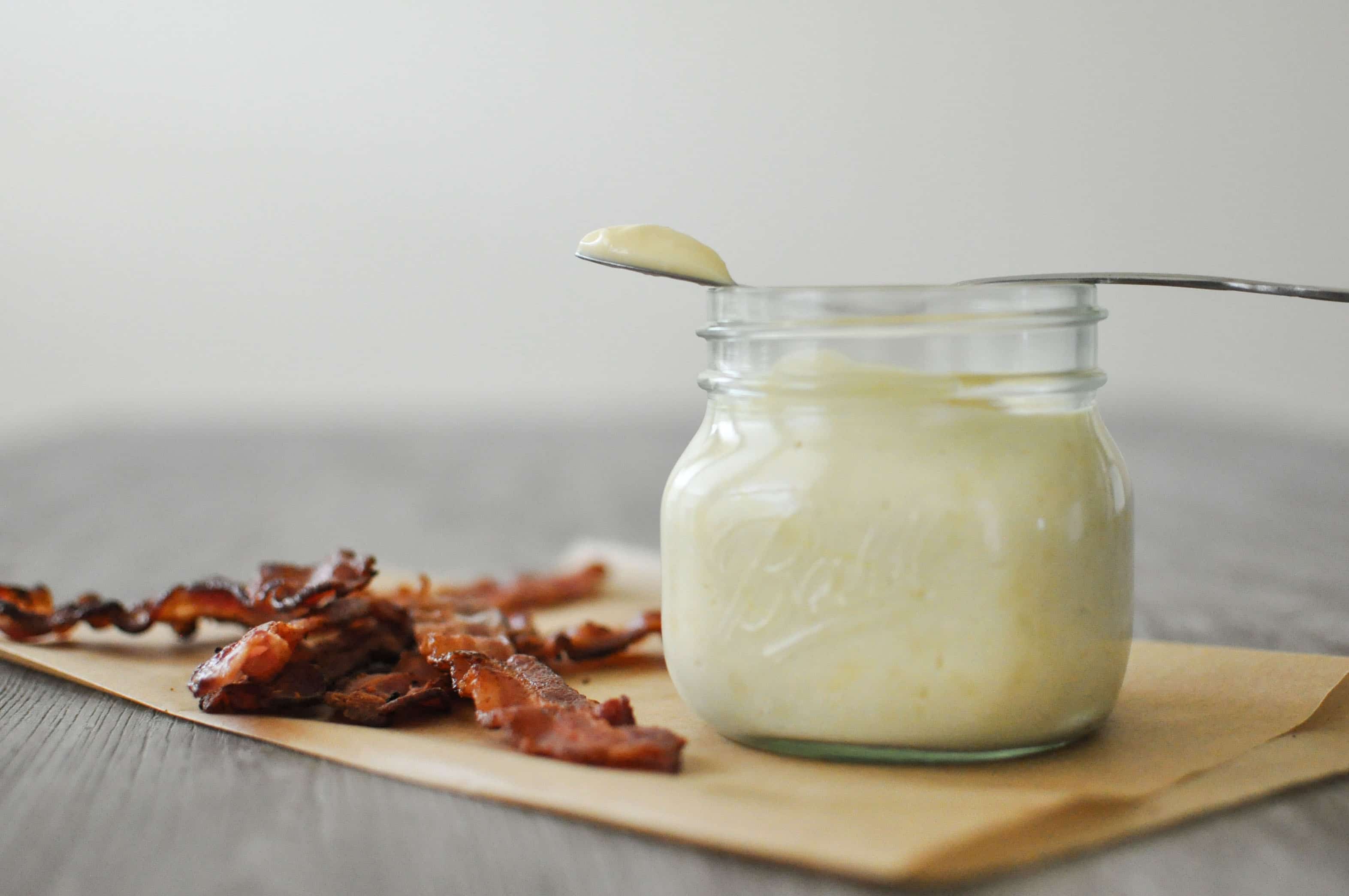 a glass jar of baconnaise on top of parchment paper next to cooked bacon with a spoon on top with a dollop of baconnaise on it