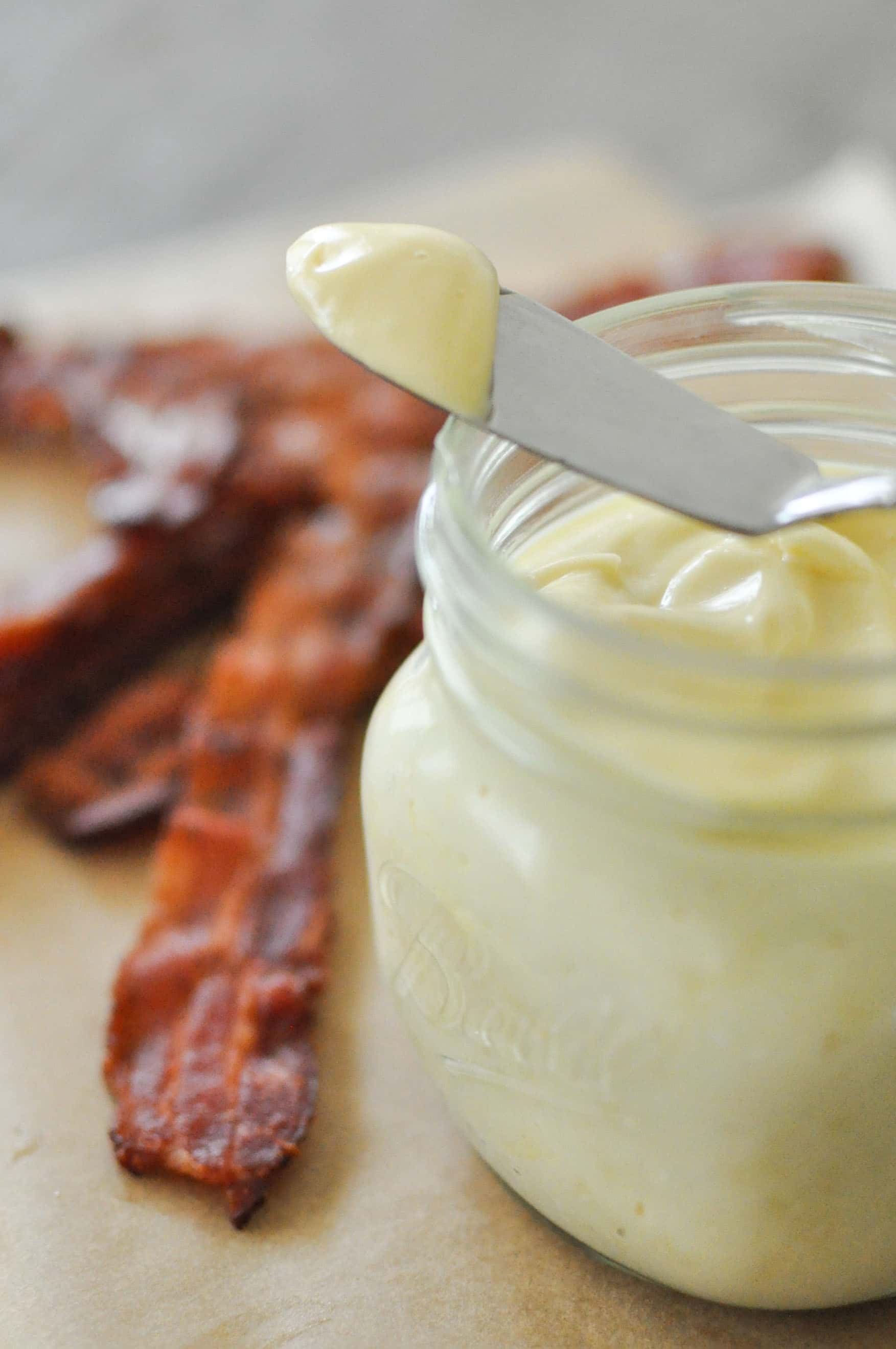 a glass jar of baconnaise on top of parchment paper next to cooked bacon with a spoon on top with a dollop of baconnaise on it