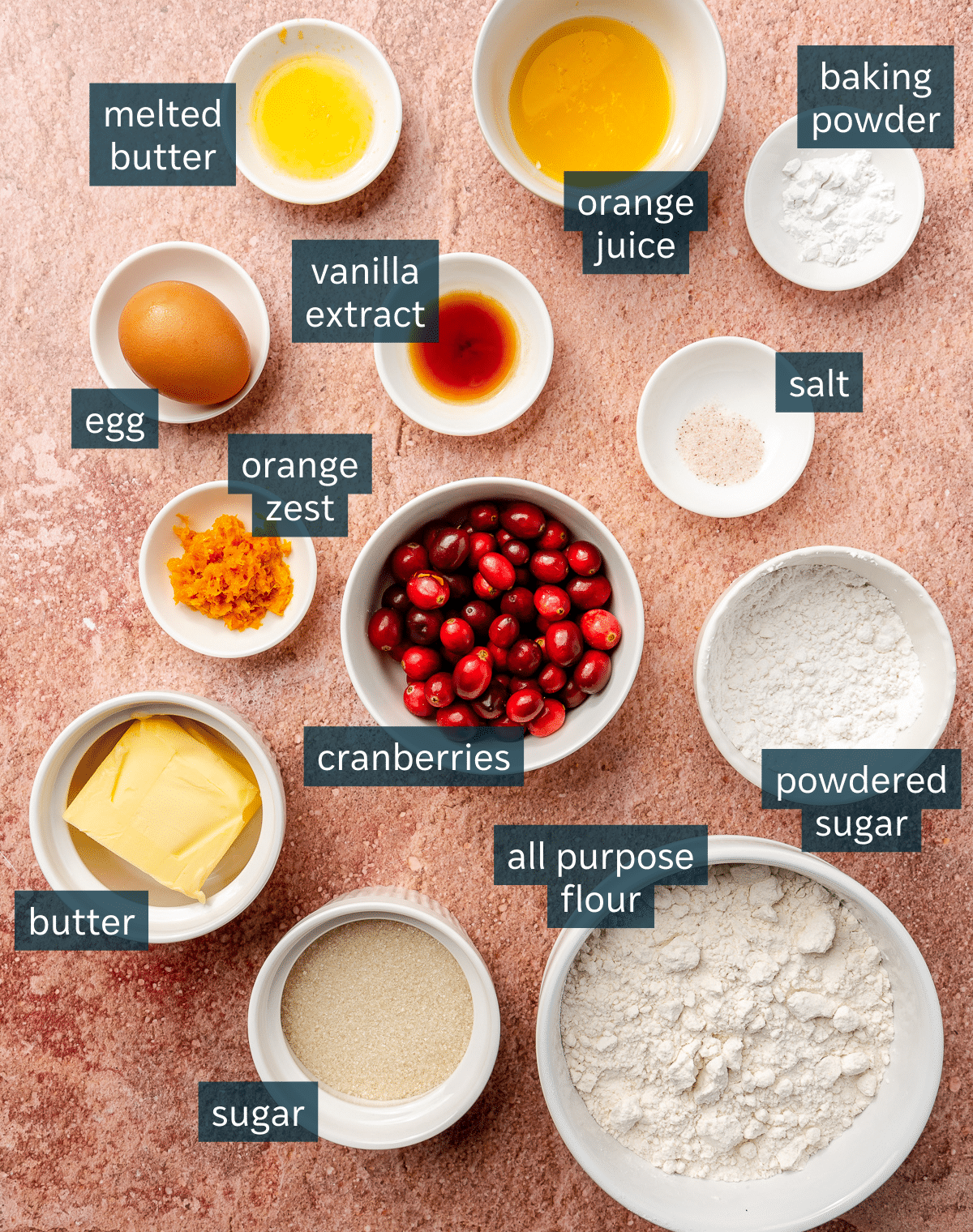 Ingredients for cranberry orange scones sit in a variety of white bowls on a light pink countertop.