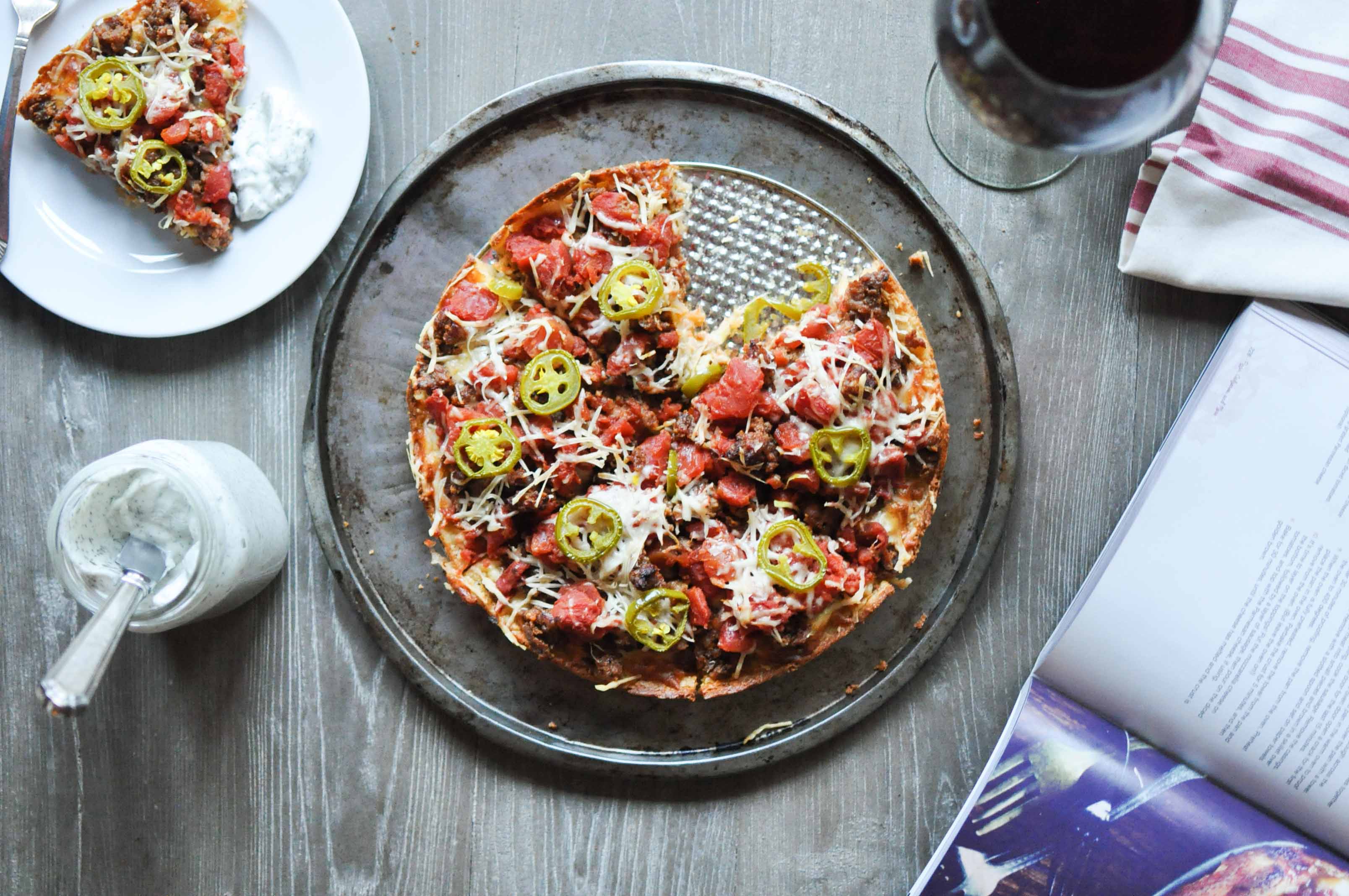 overhead view of a Chicago deep dish pizza on a round sheet pan with a plate of one slice of pizza, a jar of ranch, a glass of wine, and a book