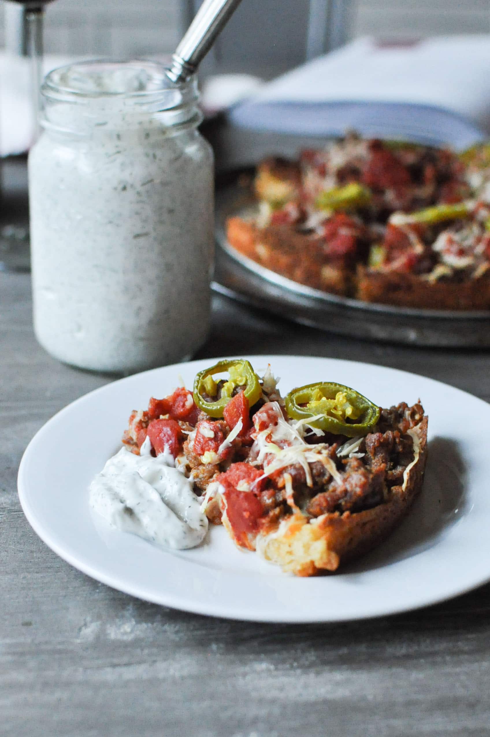 a slice of Chicago deep dish pizza on a white plate with a jar of ranch dressing and a whole pizza in the background