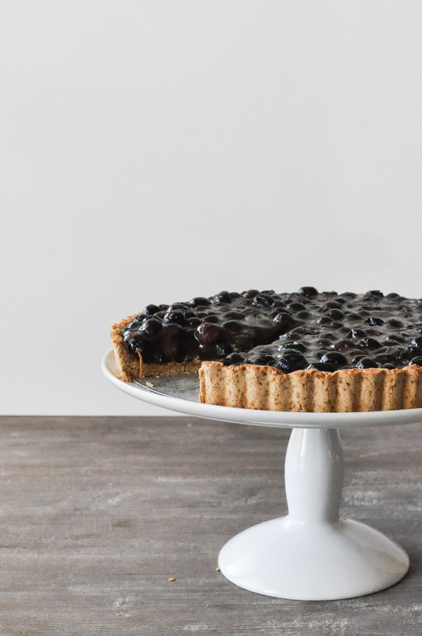 a blueberry tart on a white cake stand with one slice cut out