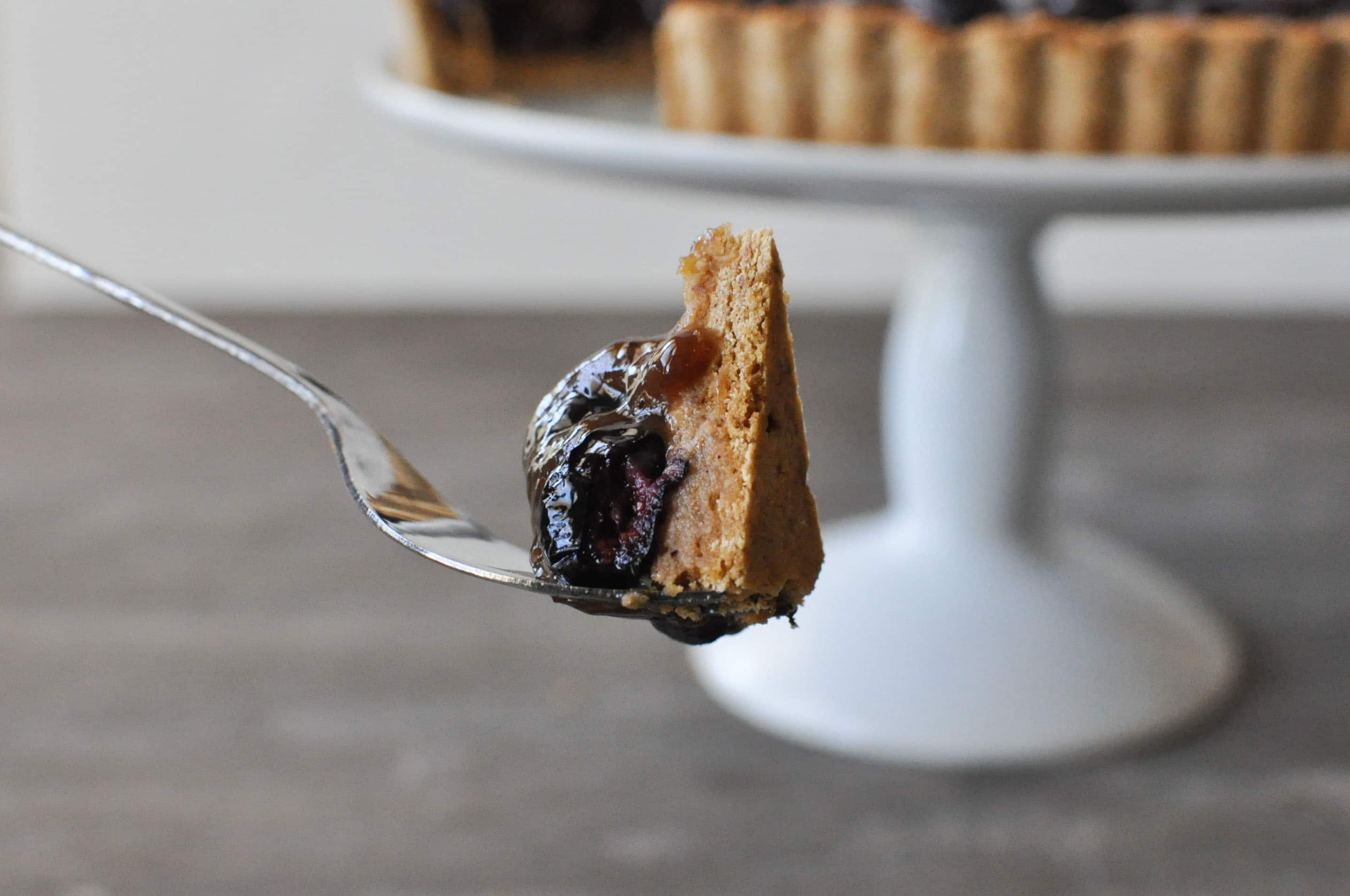 a fork holding one bite of blueberry tart with the remaining tart on a white cake stand in the background