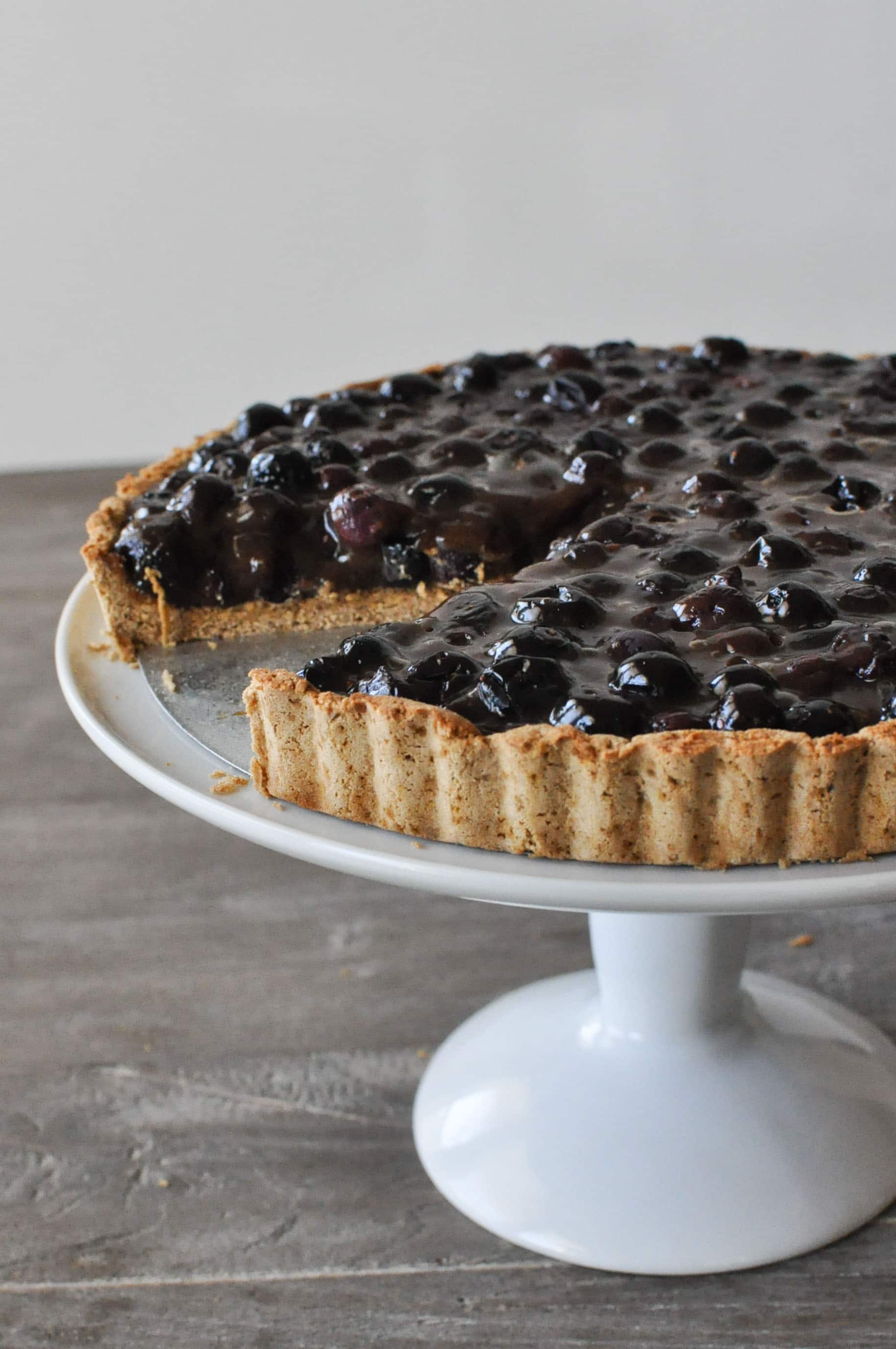 a blueberry tart on a white cake stand with one slice cut out