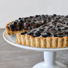 a blueberry tart on a white cake stand with one slice cut out