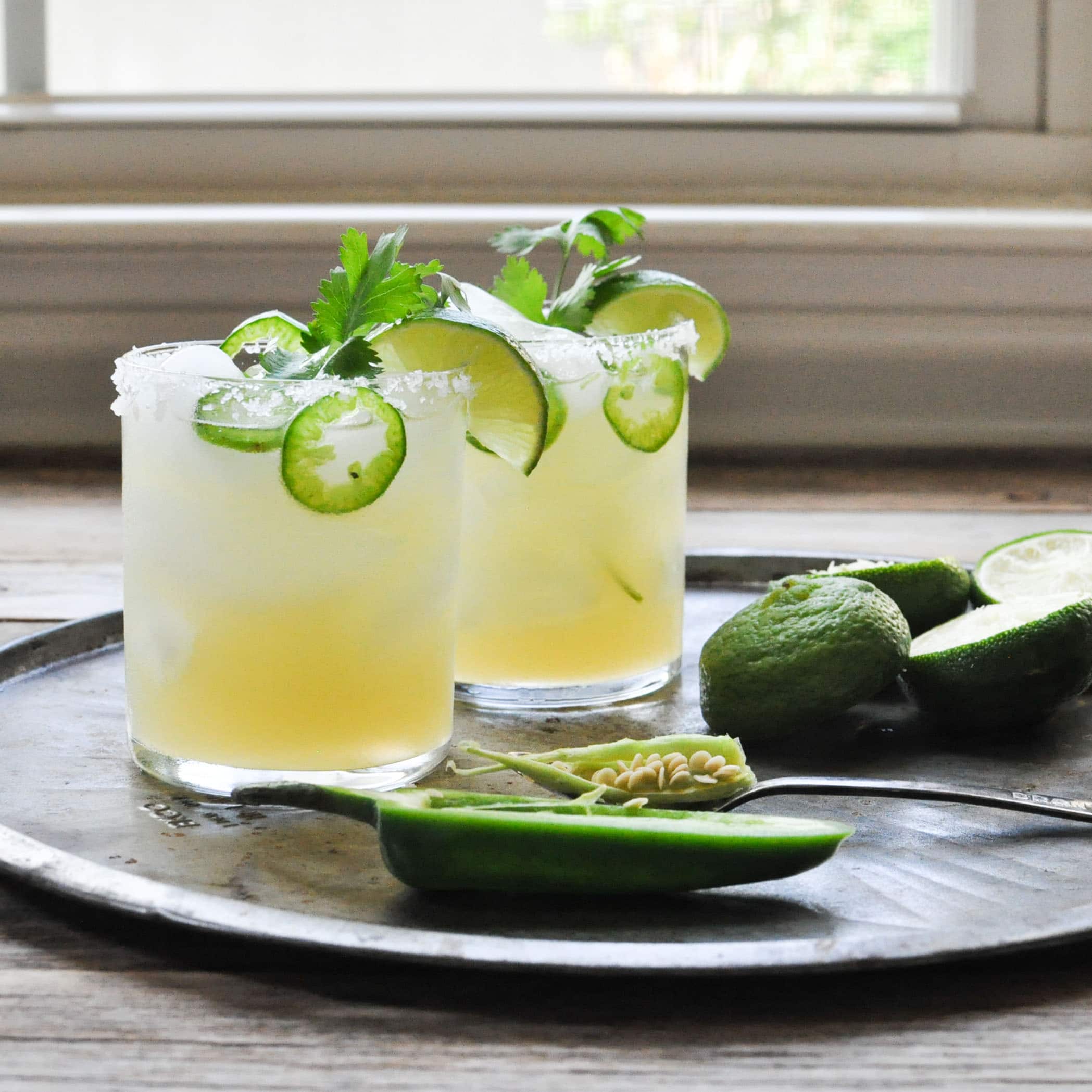 two margarita glasses filled with a fresh jalapeno and lime margaritas and garnished with a salt rim, limes, and cilantro