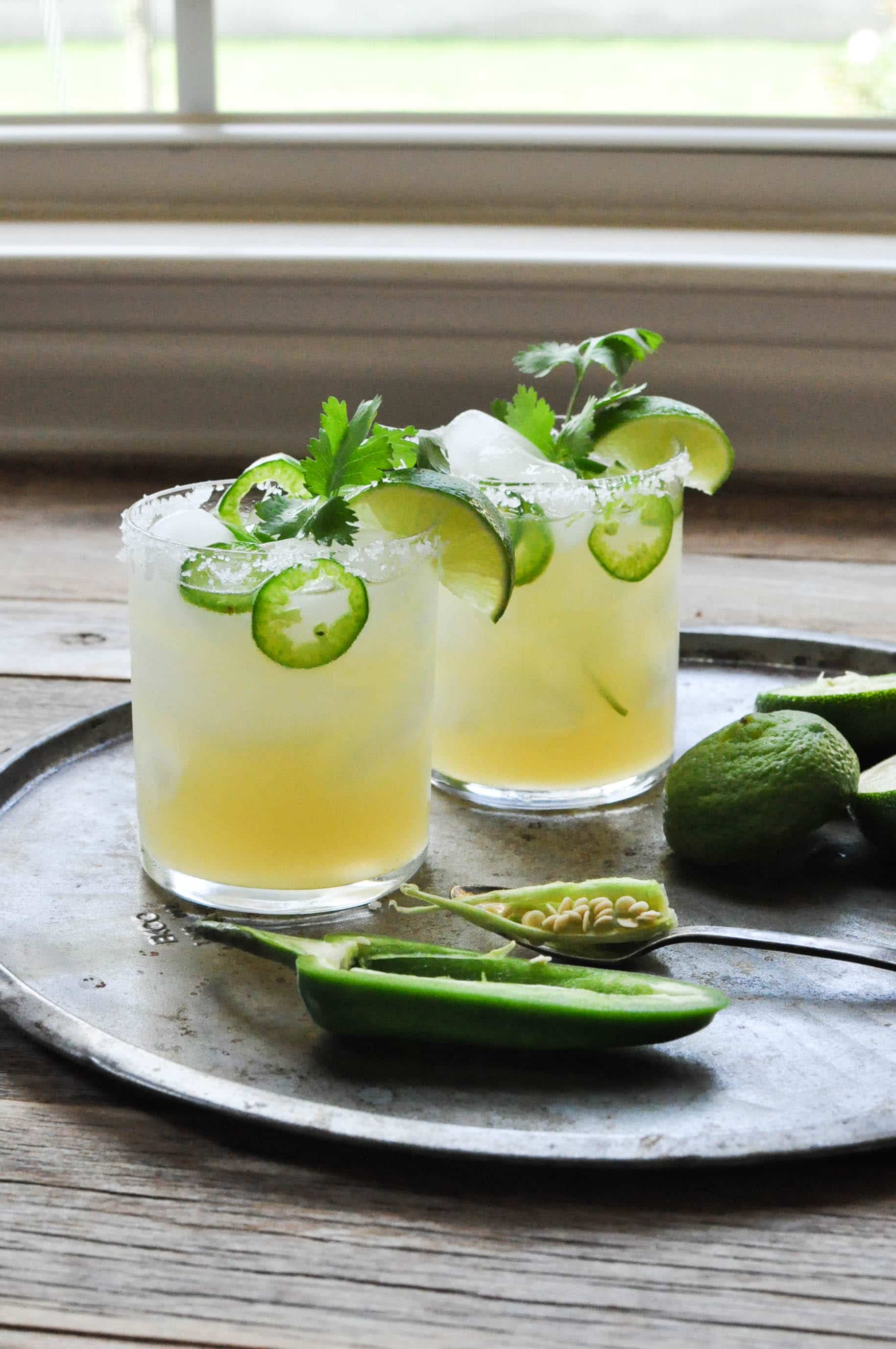 two margaritas in a glass with fresh jalapeno and lime slices on a metal place with more jalapenos and limes beside it