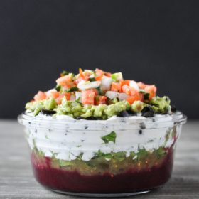 glass bowl filled with 7 layer dip and topped with pico de Gallo on a wood table