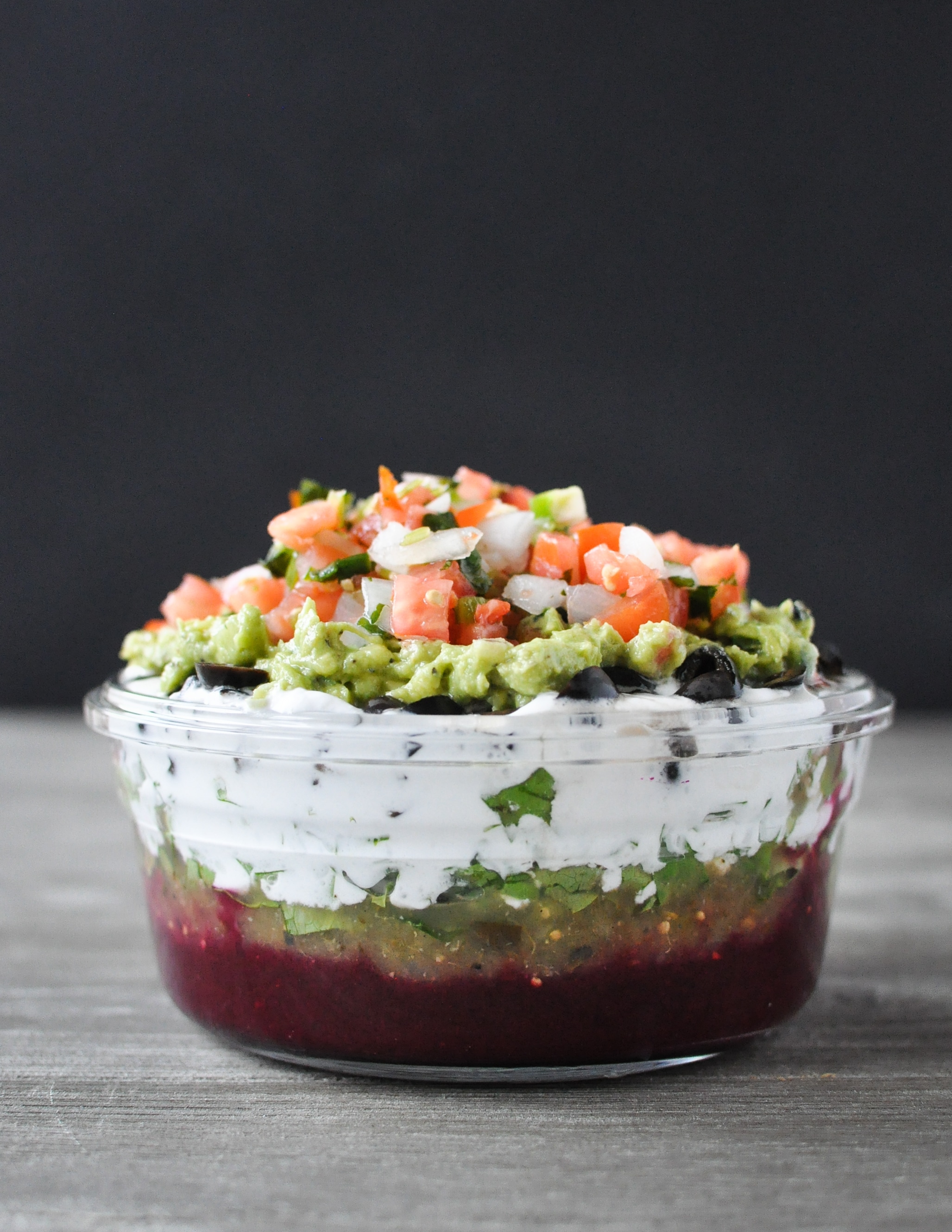 glass bowl filled with 7 layer dip and topped with pico de Gallo on a wood table