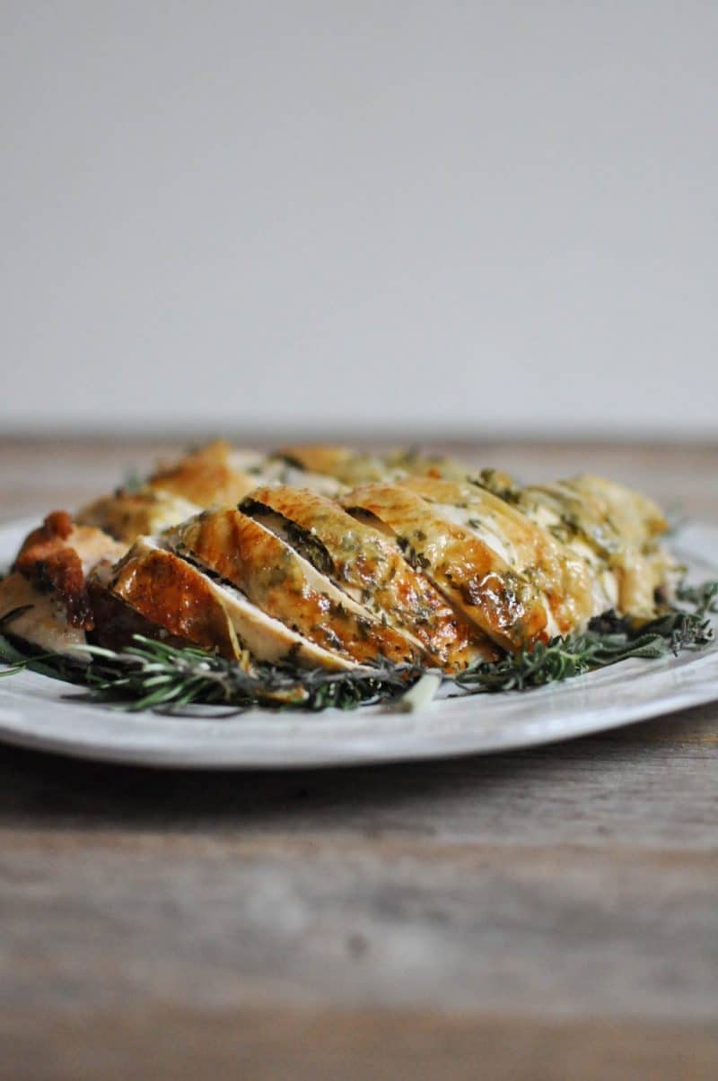 side view of sliced turkey breast with skin on a bed of herbs on a white plate on a wooden surface