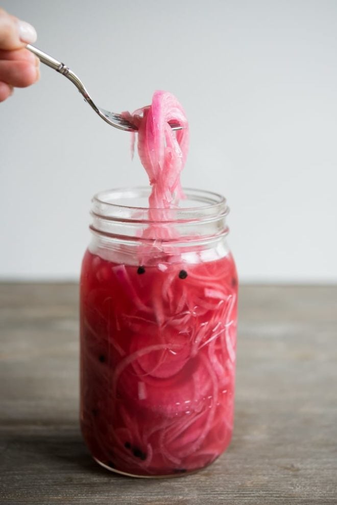 a mason jar full of pink pickled red onions on a wooden table with a fork holding some onions 