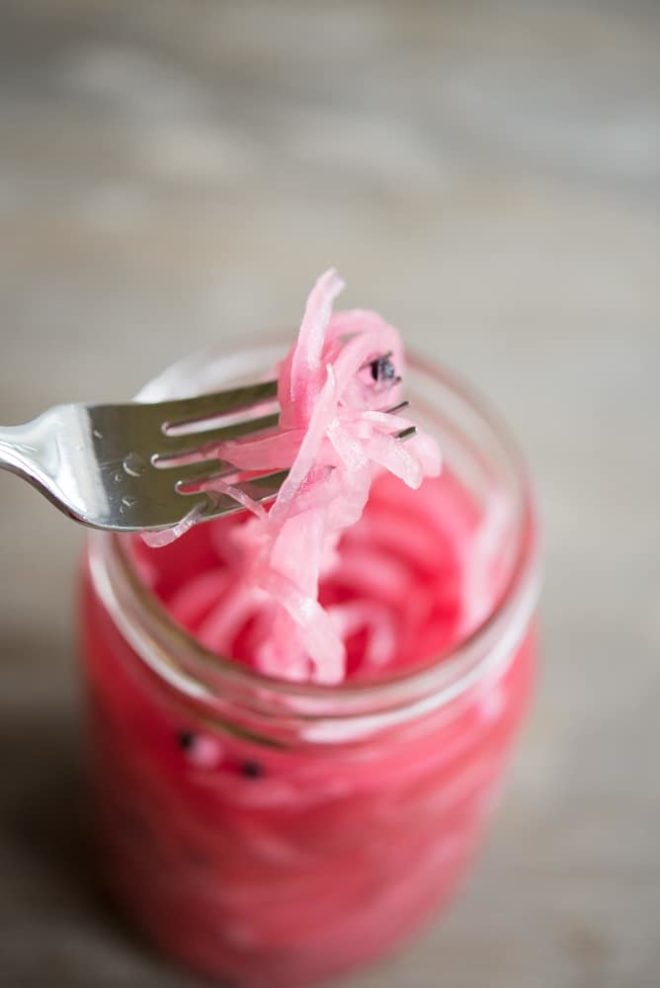 overhead view of a mason jar full of pink pickled red onions on a wooden table with a fork holding some onions