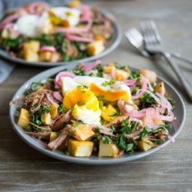 two plates loaded with potatoes, eggs, pickled onions, and corned beef (corned beef hash) sitting on a wood surface next to 2 forks