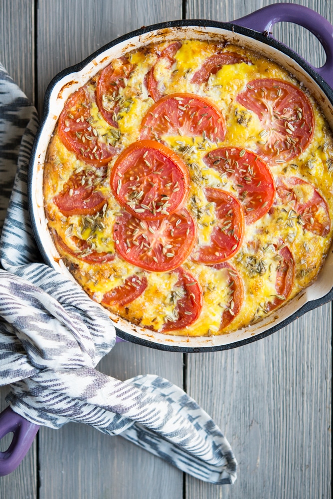 frittata topped with tomato slices in a purple enameled cast iron skillet on a light wood surface