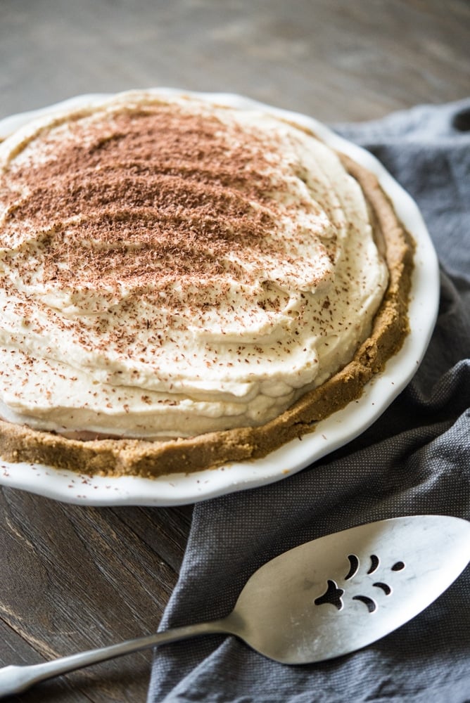 side view of chocolate mousse pie topped with whipped cream and chocolate shavings in a graham cracker crust in a white ceramic pie tin with a silver pie server and blue cloth beside it