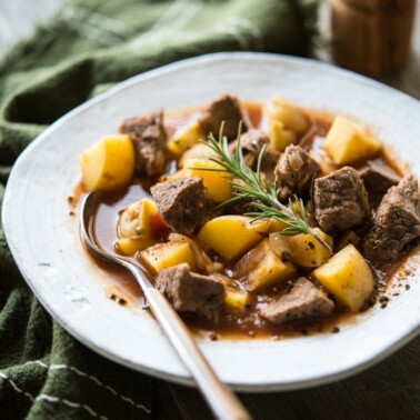 a white bowl filled with rustic beef and potato stew sitting on a green kitchen towel on a wood surface