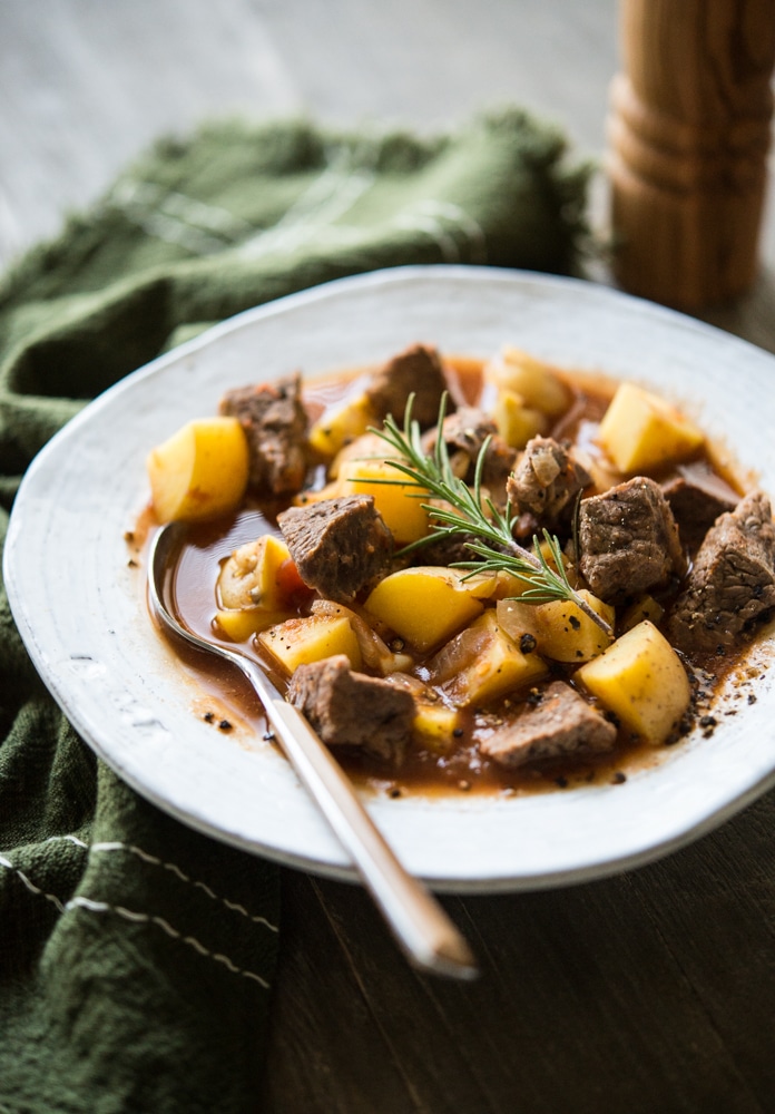 a white bowl filled with rustic beef and potato stew sitting on a green kitchen towel on a wood surface