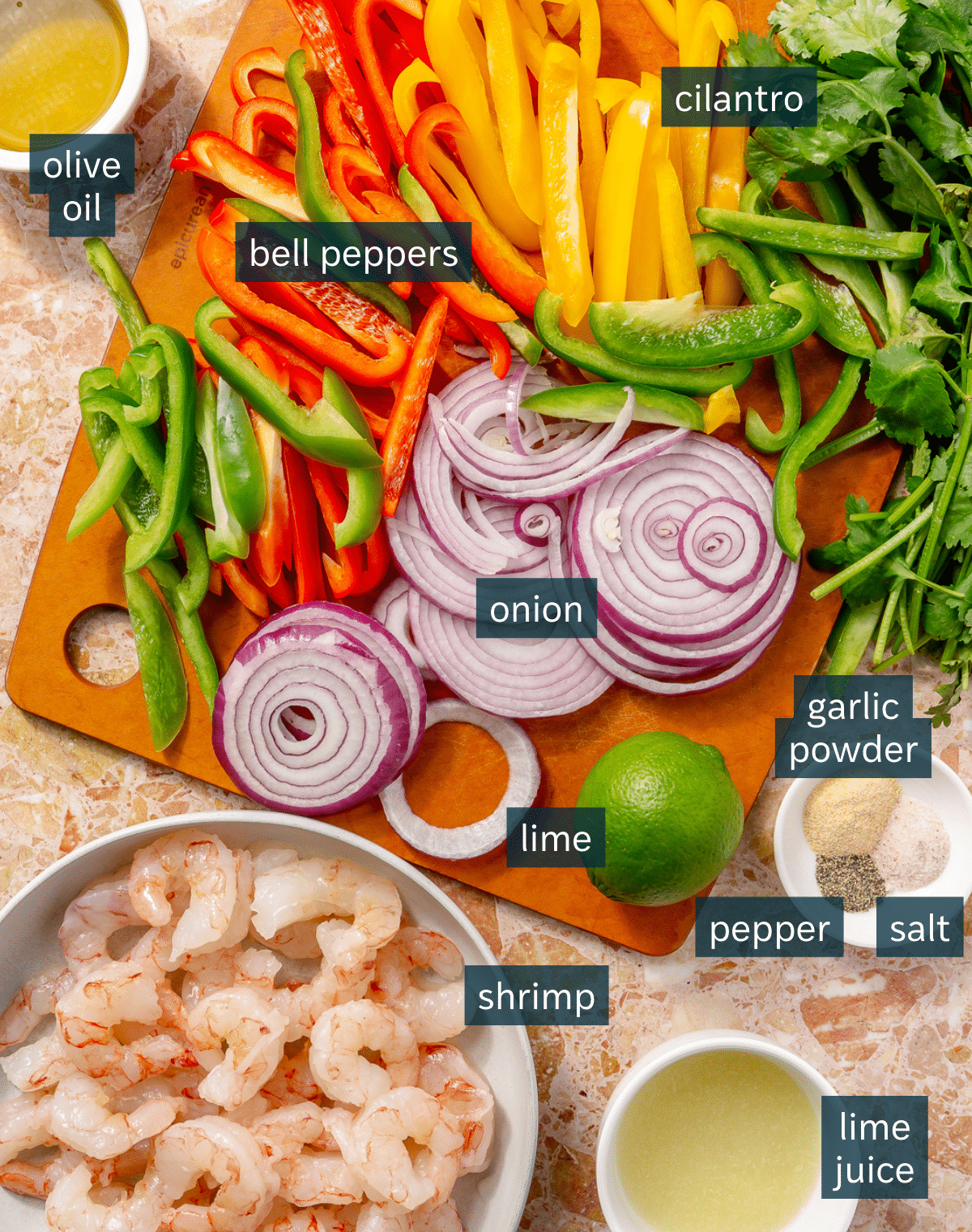 Ingredients for sheet pan shrimp fajitas sit on a cutting board and in a variety of bowls on a light pink countertop.