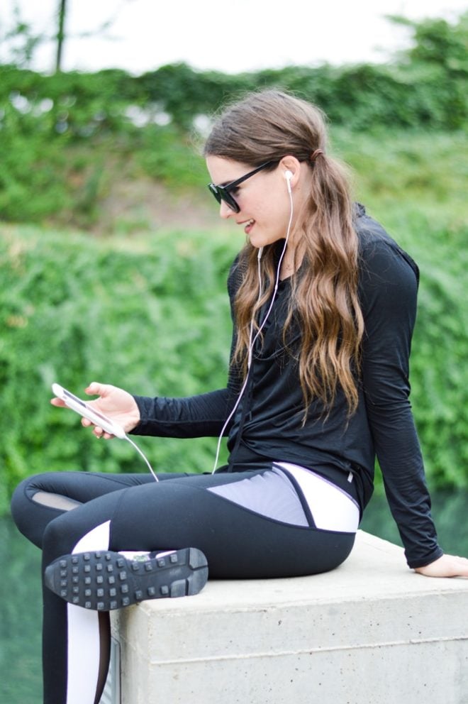 a woman with long dark pigtails sits on a concrete pillar in black activewear while wearing black sunglasses and looking down at her phone with her headphones in her ears with a green trees in the background