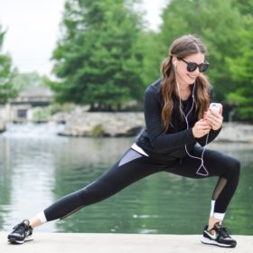 a woman with long dark pigtails lunges to the side in black activewear while wearing black sunglasses and looking down at her phone with her headphones in her ears with a green river in the background