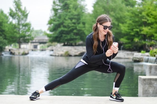 a woman with long dark pigtails lunges to the side in black activewear while wearing black sunglasses and looking down at her phone with her headphones in her ears with a green river in the background