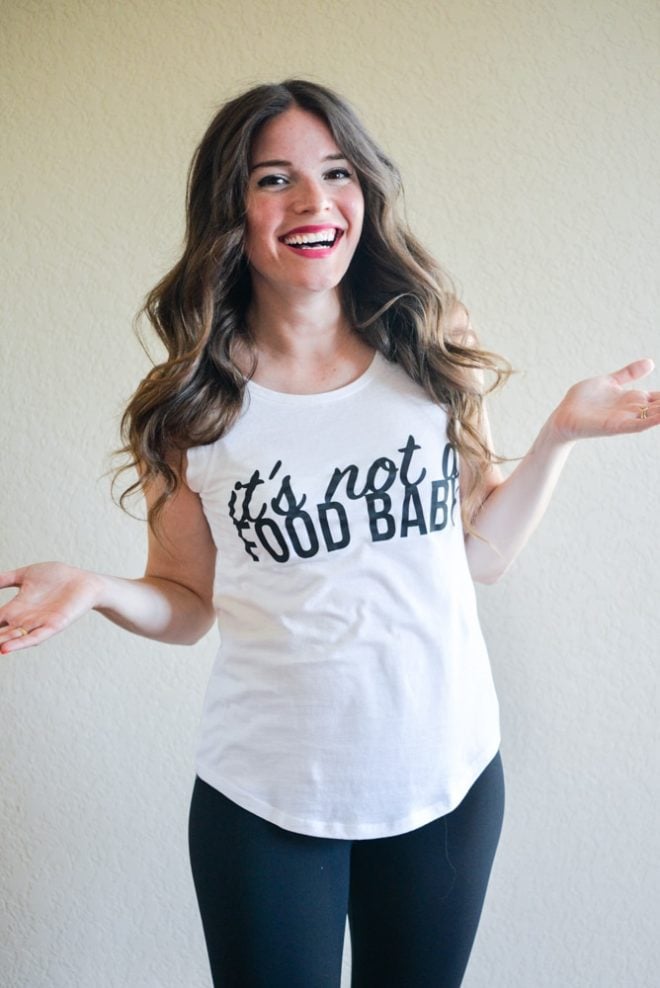 a smiling woman with long dark wavy hair stands with her hands palm up with a white shirt on that read "it's not a food baby"