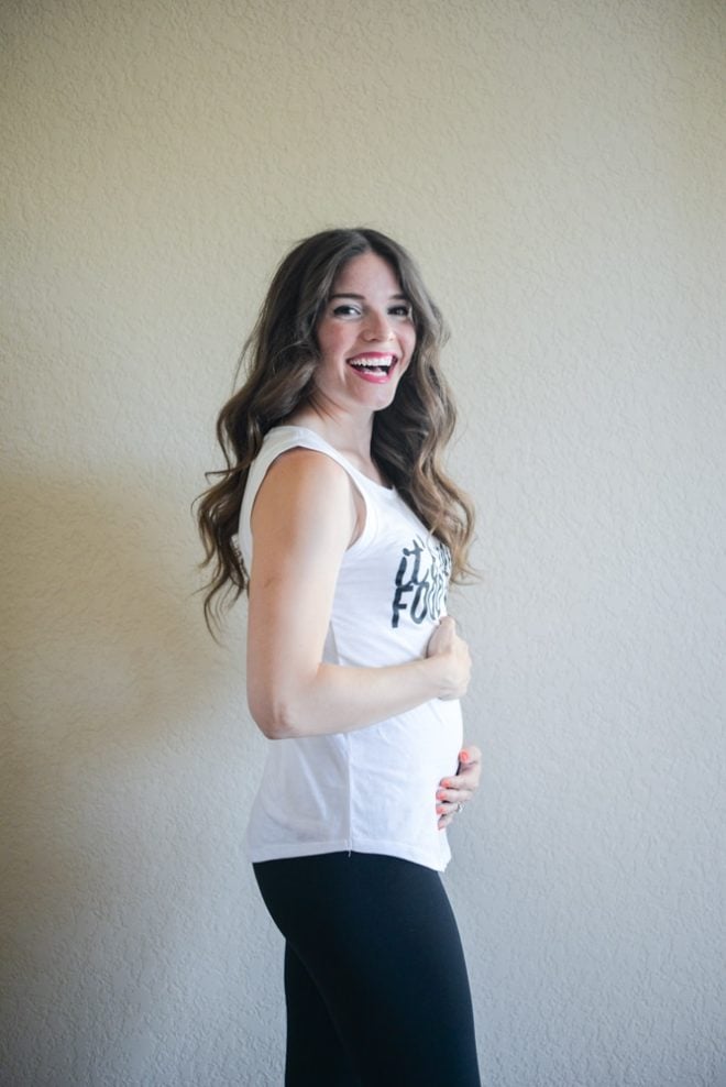 a smiling woman with long dark wavy hair stands sideways holding her newly pregnant belly