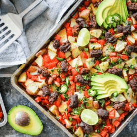 colorful potato hash with red bell peppers, chorizo, potatoes, sliced avocado, and lime wedges on a stainless steel baking sheet on a dark grey surface with a grey towel and spatula alongside it