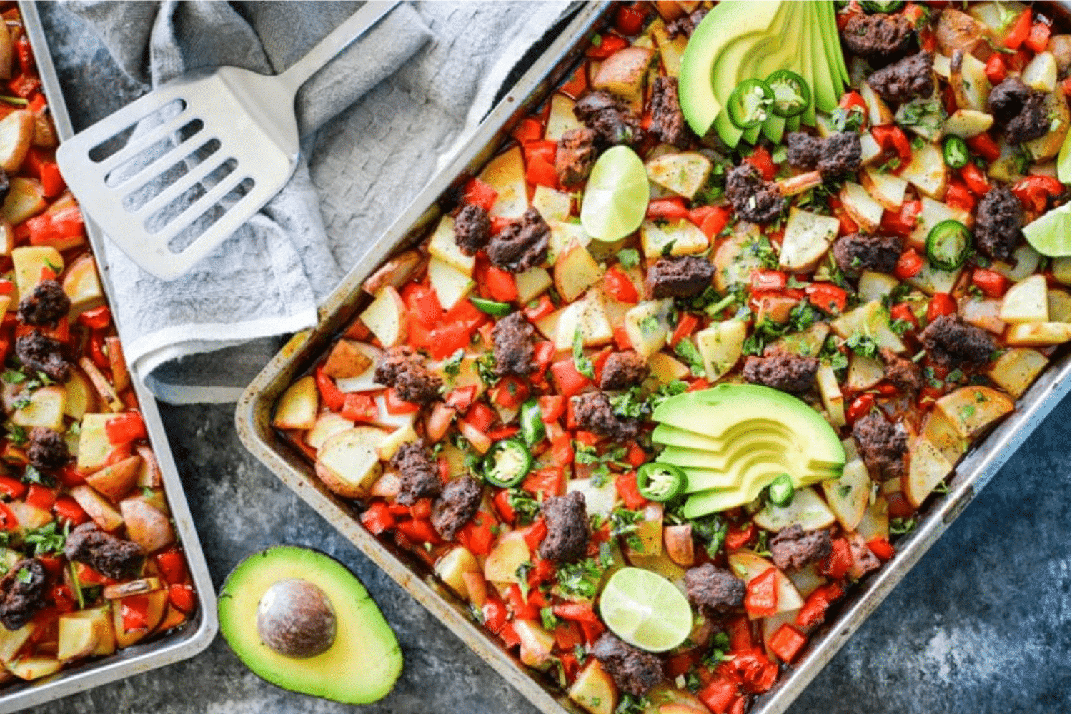 colorful potato hash with red bell peppers, chorizo, potatoes, sliced avocado, and lime wedges on a stainless steel baking sheet on a dark grey surface with a grey towel and spatula alongside it