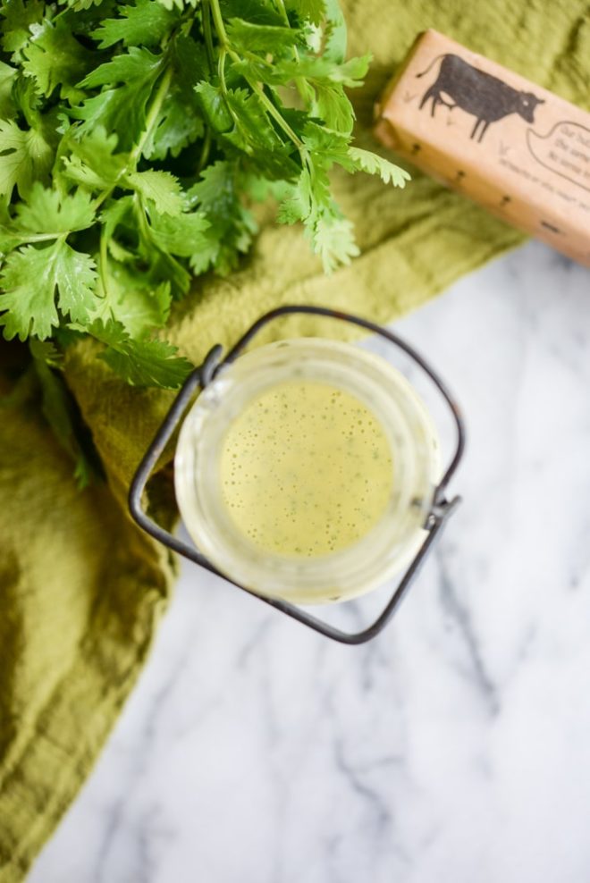overhead view of a glass jar of cilantro hollandaise sauce on a white marble table with cilantro in the background