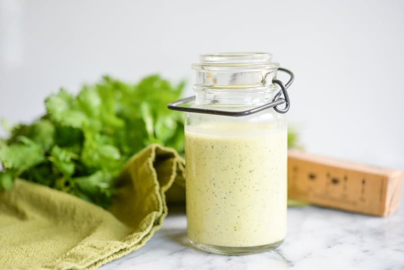 glass jar of cilantro hollandaise sauce on a white marble table with cilantro in the background