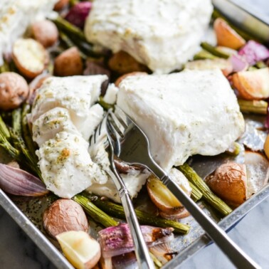 baked halibut filets on top of roasted potatoes, red onions, and asparagus on a sheet pan with two forks digging into the halibut