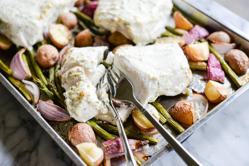 baked halibut filets on top of roasted potatoes, red onions, and asparagus on a sheet pan with two forks digging into the halibut