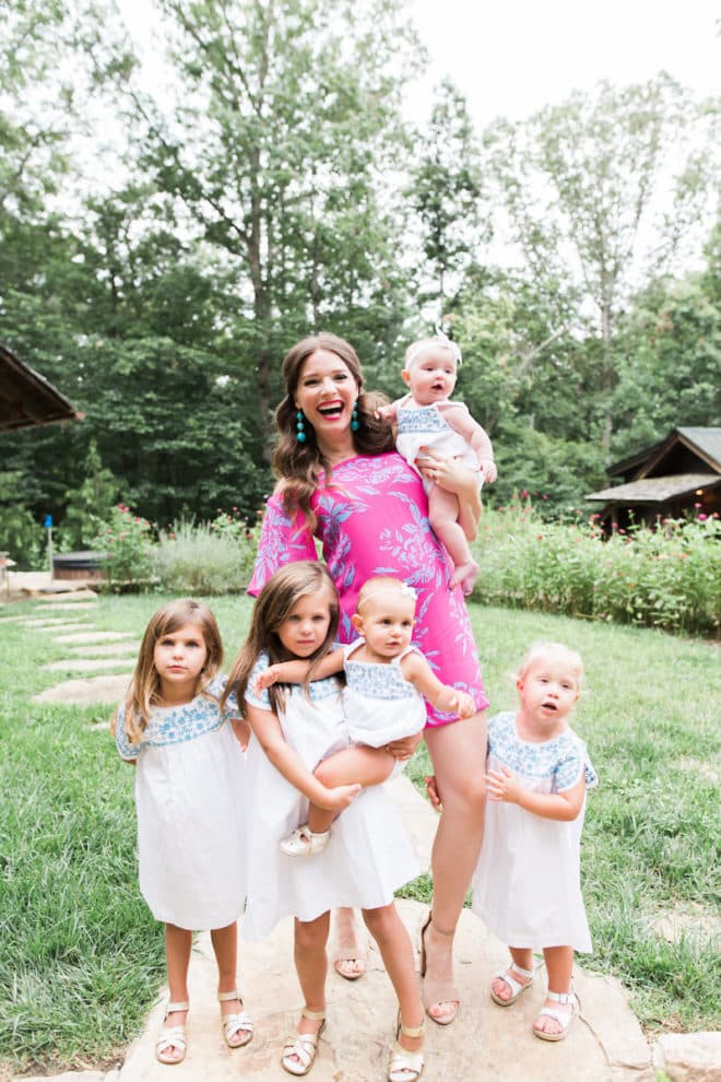 a long dark haired pregnant women in a bright pink floral dress holds a baby while 4 children pose in front of her in white and blue dresses in a green wooded area