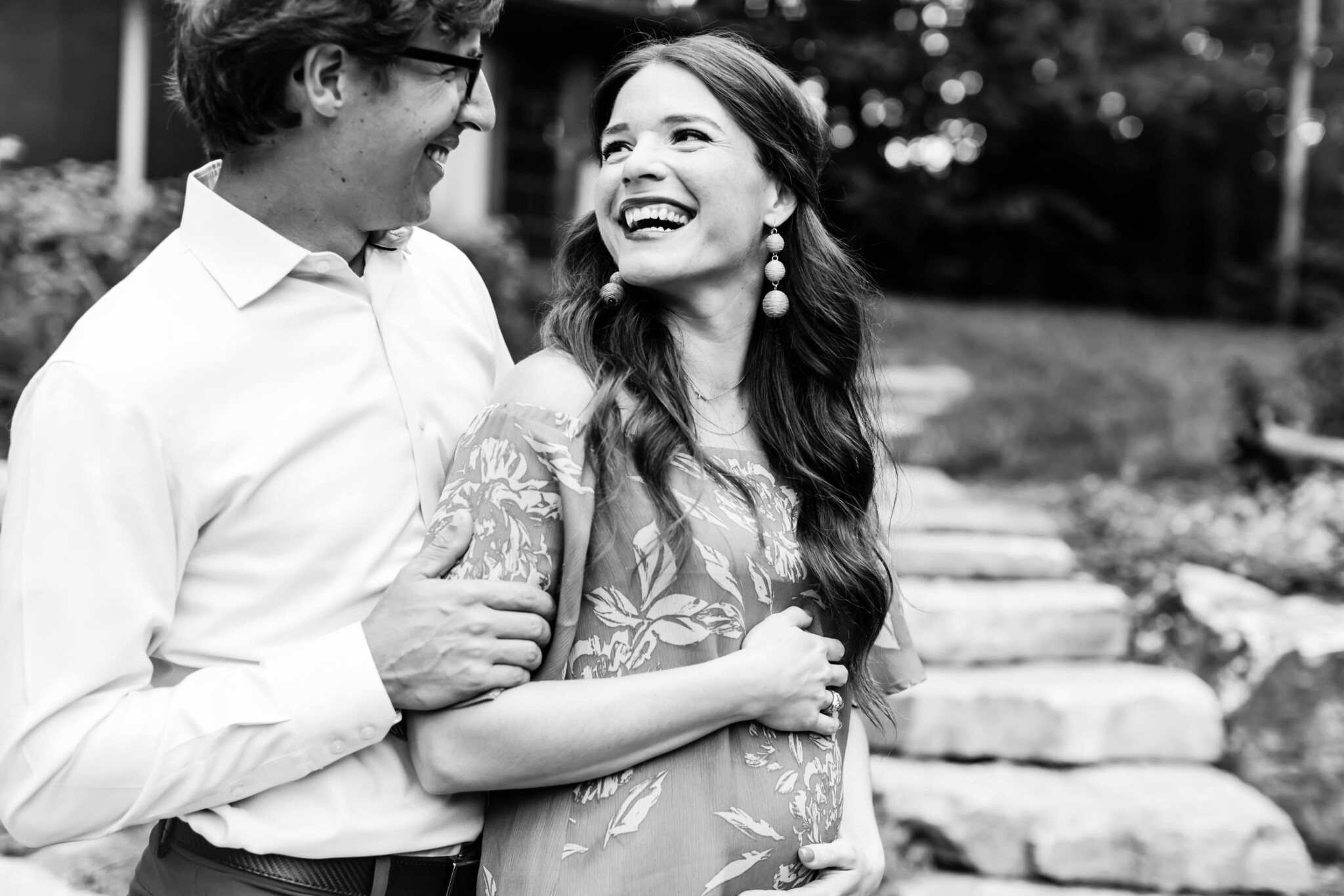 black and white photo of a man in a white button down collared shirt in black glasses holding his long dark haired white who is looking back at him and smiling while cradling her pregnant belly