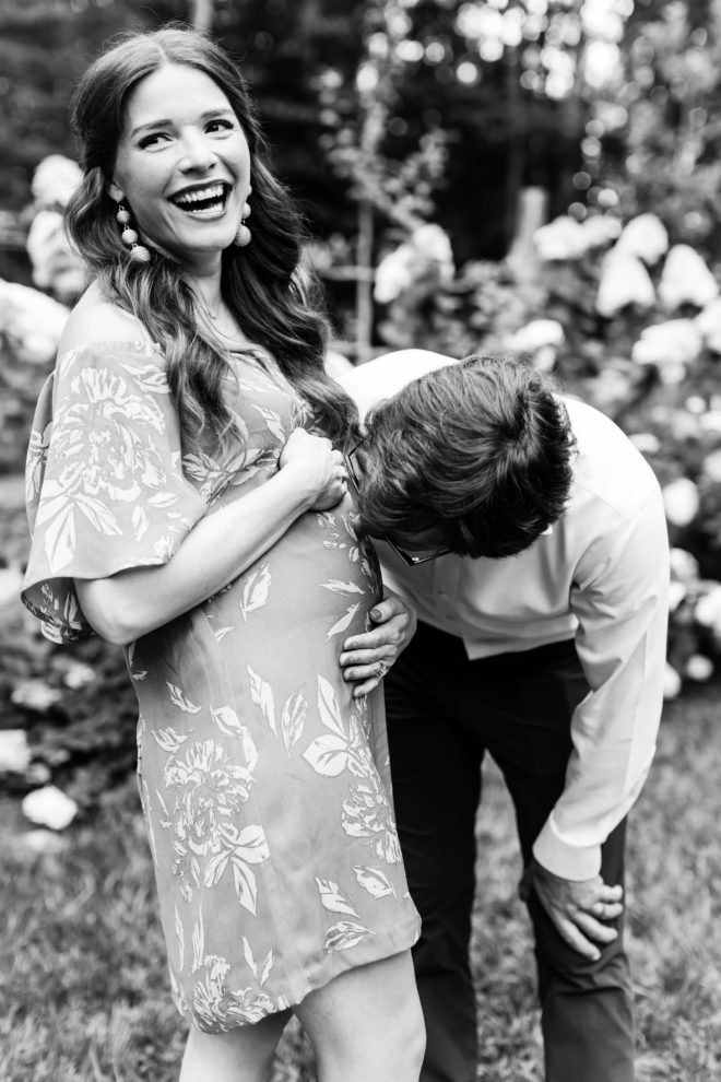 black and white image of a long dark haired smiling woman cradling her pregnant belly with her husband leaning down to kiss it