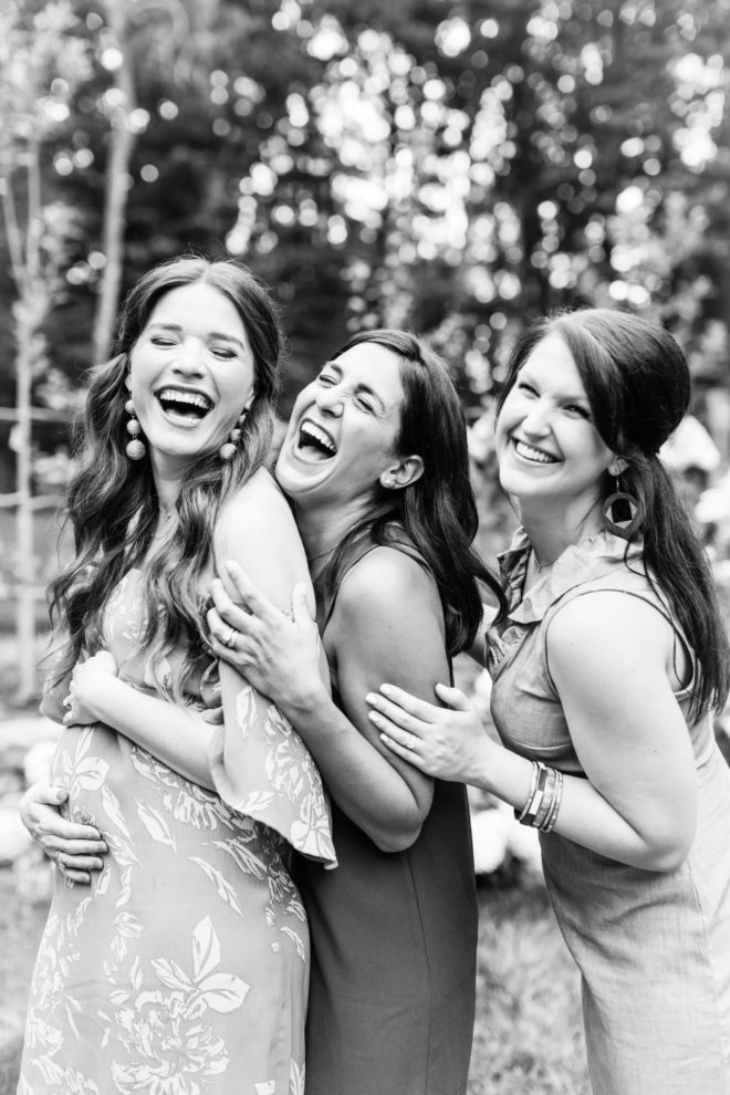 black and white image of three smiling happy women with long dark hair holding each other and one cradling her pregnant belly