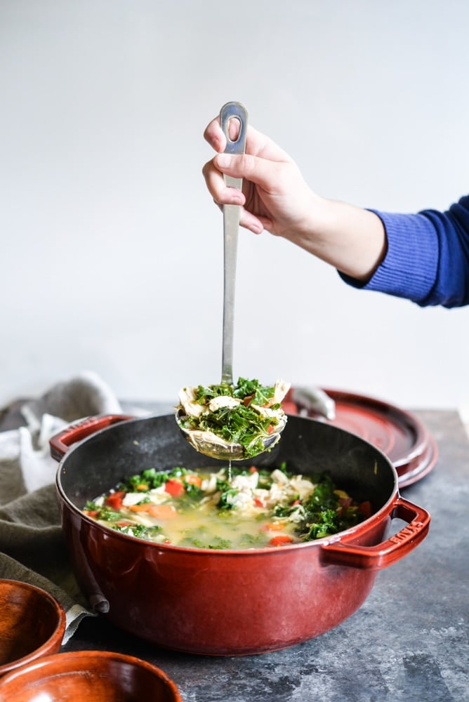 chicken soup in a ladle being held over a large red pot filled with soup