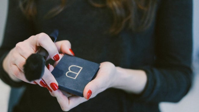 a woman in a black shirt with long dark hair and red fingernails holding a blush palette and a blush brush 